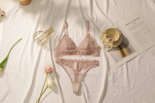 A delicate lace bra set in pink, displayed on a flat surface with a book, gold-colored brassiere, and floral arrangement in the background.